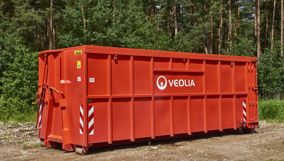 Ein großer, roter Veolia-Abrollcontainer steht auf einem unbefestigtem Boden. Im Hintergrund sieht man einen dichten, grünen Wald.