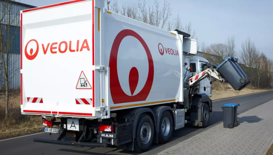 Ein weißer Seitenlader mit Veolia Logo der am Straßenrand steht. Das Fahrzeug hebt eine schwarze Mülltonne an.