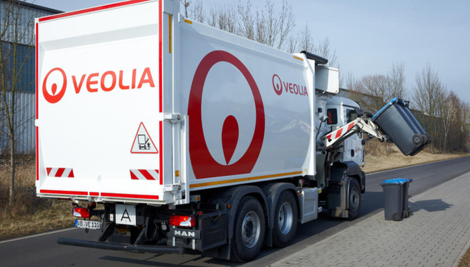 Ein weißer Seitenlader mit Veolia Logo der am Straßenrand steht. Das Fahrzeug hebt eine schwarze Mülltonne an.
