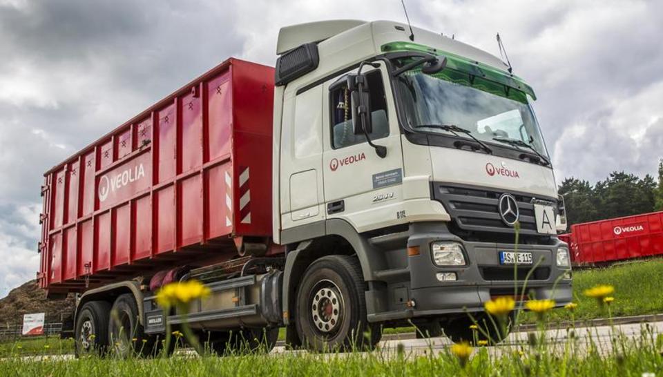 Ein weißer Abrollkipper mit Veolia-Logo der auf einer Straße fährt. Der Container, mit dem er beladen ist, hat eine leuchtend rote Farbe. Im Vordergrund des Bildes sind leicht verschwommene Gräser wahrzunehmen. 
