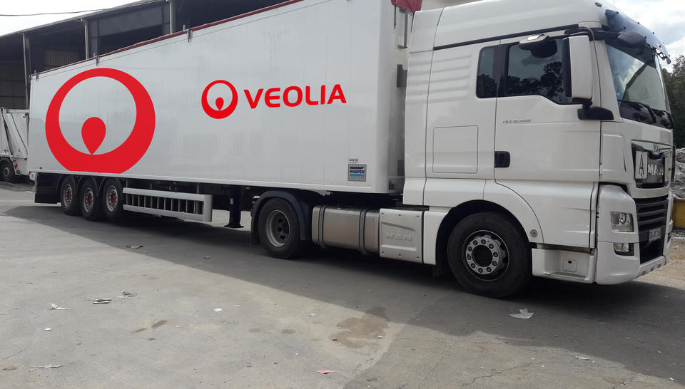 Das Bild zeigt einen weißen LKW mit rotem Veolia-Logo. Es handelt sich um einen Sattelschlepper mit Auflieger. Der LKW steht auf einer betonierten Fläche, dem Betriebshof von Veolia.