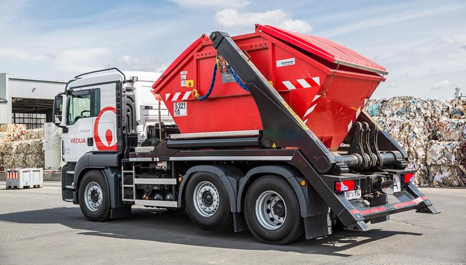 Ein weißer Veolia-LKW, ein sogenannter Absetzkipper. Der mit einem rot leuchtendem Container beladen ist. 