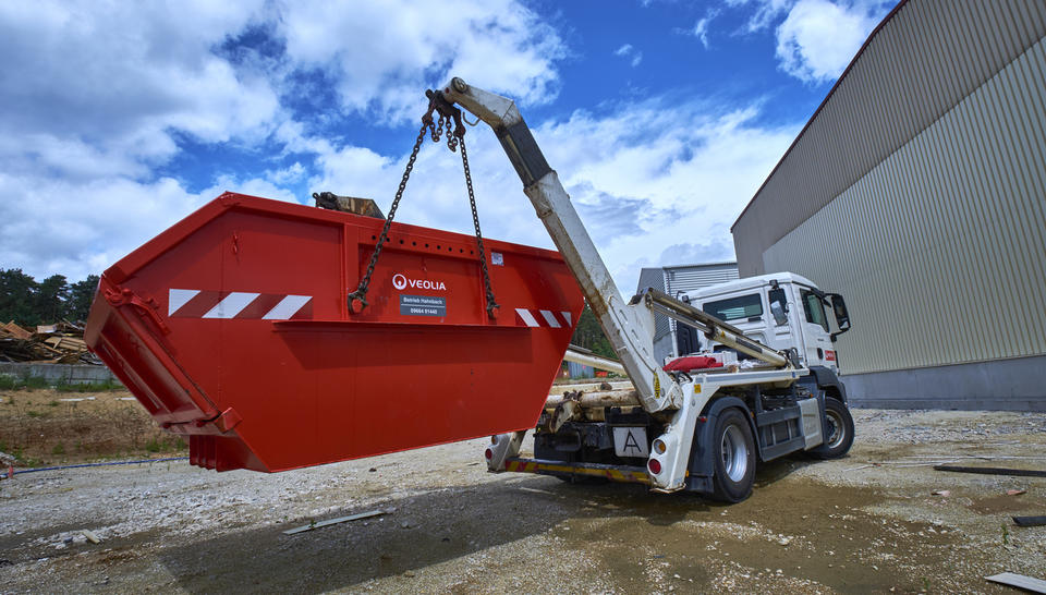 "Ein LKW mit Hebevorrichtung hebt einen großen roten Absetzcontainer der Firma Veolia an. Der Container wird von Ketten in der Luft gehalten, die an einem weißen Kranarm befestigt sind. Der Kranarm gehört zu dem weißen LKW. "