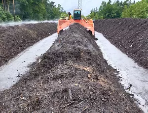 Frontalaufnahme von drei Reihen Kompost. Im Hintergrund der mittleren Reihe der Komposte ist vermutlich ein orangenfarbener Kompostwender zu sehen. 