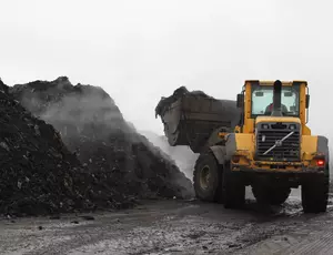 Im Bild sind auf der linken Seite zwei dunkle Hügel mit Klärschlamm zu sehen. Rechts davon, steht ein gelber Radlader der erneut Klärschlamm auf einem der Hügel verteilt. 