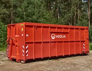 Ein großer, roter Veolia-Abrollcontainer steht auf einem unbefestigtem Boden. Im Hintergrund sieht man einen dichten, grünen Wald.