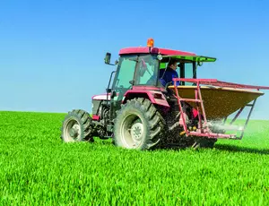 Ein roter Traktor auf einem grünen Feld. Der Traktor streut Dünger. Im Hintergrund strahlt der blaue Himmel.