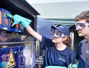 Zwei junge Personen in blauen T-Shirts und Schutzbrillen stehen vor einem geöffneten Schaltschrank. Eine Person greift in den Schrank, während die andere zuschaut. Der Schrank enthält verschiedene elektrische Komponenten und Warnzeichen.