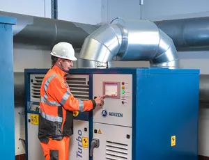 Ein Mitarbeiter in orangefarbener Sicherheitskleidung und einem Sicherheitshelm, bedient eine Belüftungsanlage des Klärwerks in Schönebeck. Im Hintergrund befinden sich 2 große, graue Lüftungsrohre.