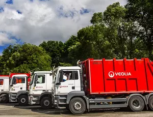 Mehrere Müllfahrzeuge mit Veolia-Logo stehen hintereinander auf einem Parkplatz. Im Hintergrund sind viele grüne Bäume und der Himmel mit Wolken zu sehen.