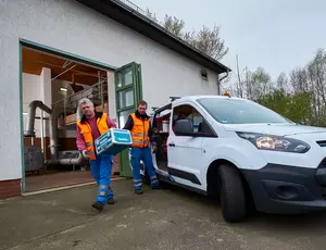 Zwei Menschen in Sicherheitskleidung laden etwas in einen weißen Lieferwagen. Im Hintergrund ist ein weißes Gebäude mit geöffnetem Tor zu erkennen.