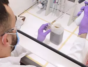 Nahaufnahme eines Labortisches. Eine Person in Schutzkleidung, mit Maske und lila Handschuhen, führt eine Wasserprobe durch. Zu sehen sind Laborgeräte wie Glasgefäße und Pipetten. 