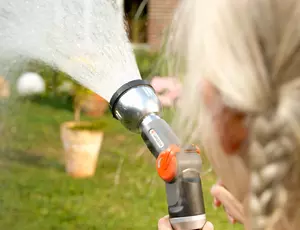 Aufnahme von hinten: Auschnitt eines Mädchens mit blonden Haaren und langen Zöpfen, die eine Wasserschlauchpistole in der Hand hält. Im Hintergrund ist unscharf grünes Gras zu erkennen.