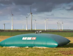 Im Vordergrund befindet sich ein großer, aufblasbarer Wasserspeicher, eine sogenannte Löschwasserzisterne. Dahinter ist ein Windpark mit mehreren Windrädern. Darüber der bewölkte Himmel.