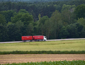 Luftaufnahme eines fahrenden weißen Veolia-LKW mit zwei roten Anhängern. Der LKW fährt auf einer Straße. Auf der linken Seite der Straße befindet sich ein Wald, auf der rechten ein Feld.
