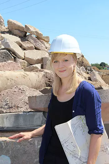 Person mit weißem Schutzhelm auf einem Baustellen- oder Recyclinggelände. Die Person hält Baupläne oder technische Zeichnungen in der Hand und steht vor einem Hintergrund aus großen Beton- und Gesteinsbrocken. Das Bild vermittelt die Planung, Überwachung oder Leitung von Bau-, Umwelt- oder Entsorgungsprojekten.