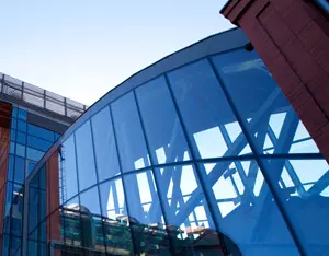 Auf dem Bild ist ein modernes Gebäude mit einer großen, gebogenen Glasfassade zu sehen. Die Fassade besteht aus zahlreichen blauen Glasscheiben, in denen sich die Umgebung und die Konstruktion des Gebäudes spiegeln. Hinter dem Glas sind tragende Stahlstreben in einer geometrischen Anordnung erkennbar. Links und rechts wird die Glasfront von roten Gebäudeteilen eingerahmt. Der klare Himmel und die moderne Architektur vermitteln einen technologischen und innovativen Eindruck.