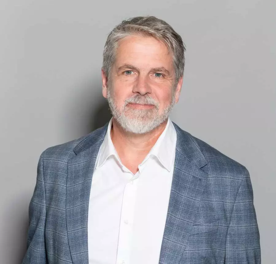 Porträtaufnahme: Ein Mann mittleren Alters mit grauen Haaren und Vollbart. Er trägt einen grauen Anzug und ein weißes Hemd.