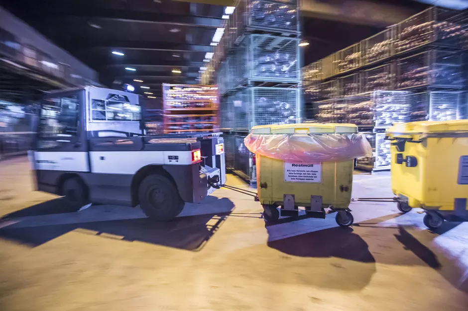 Auf dem Bild ist eine Lager- oder Industriehalle zu sehen. Im Vordergrund fährt ein kleines Transportfahrzeug zwischen mehreren großen gelben Abfallcontainern hindurch. Die Container stehen auf Rollen und sind teilweise mit Müllsäcken ausgekleidet. Im Hintergrund befinden sich gestapelte Gitterboxen und weitere Lagerflächen. Die Szene vermittelt den Eindruck einer organisierten Abfalllogistik, bei der Abfälle innerhalb einer Anlage gesammelt, transportiert und für die weitere Behandlung oder das Recycling v