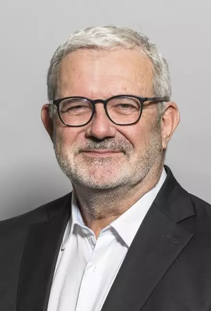 Porträtaufnahme: Ein Mann mit grauen Haaren und Vollbart.Er trägt einen dunklen Anzug und ein weißes Hemd.