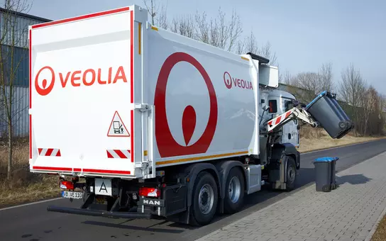 Ein weißer Seitenlader mit Veolia Logo der am Straßenrand steht. Das Fahrzeug hebt eine schwarze Mülltonne an.