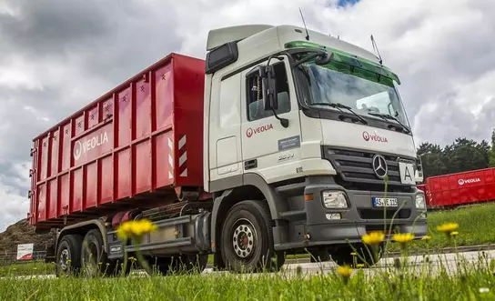 Ein weißer Abrollkipper mit Veolia-Logo der auf einer Straße fährt. Der Container, mit dem er beladen ist, hat eine leuchtend rote Farbe. Im Vordergrund des Bildes sind leicht verschwommene Gräser wahrzunehmen. 