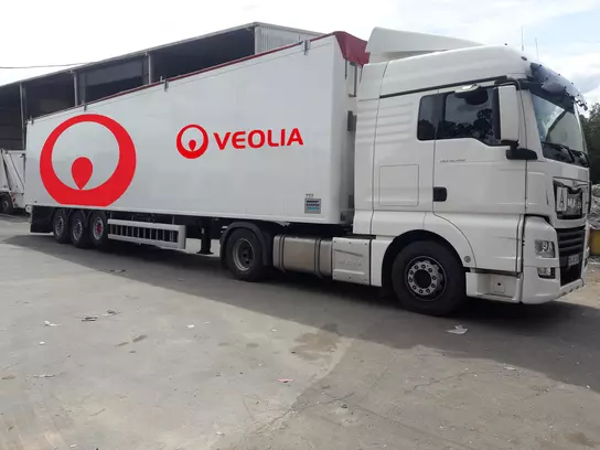 Das Bild zeigt einen weißen LKW mit rotem Veolia-Logo. Es handelt sich um einen Sattelschlepper mit Auflieger. Der LKW steht auf einer betonierten Fläche, dem Betriebshof von Veolia.