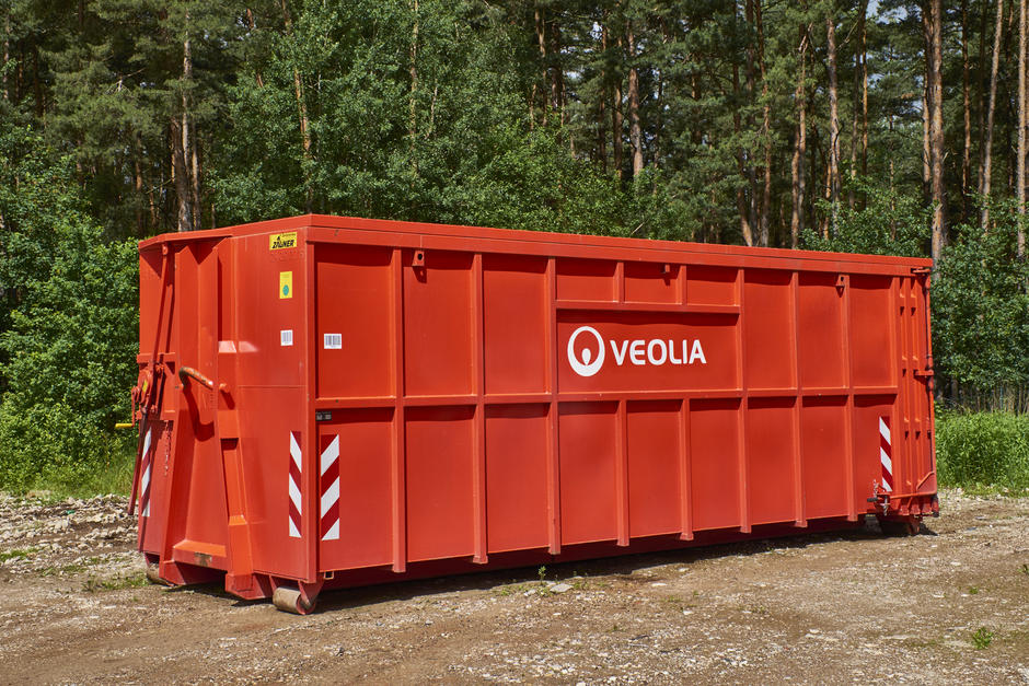 Ein großer, roter Veolia-Abrollcontainer steht auf einem unbefestigtem Boden. Im Hintergrund sieht man einen dichten, grünen Wald.