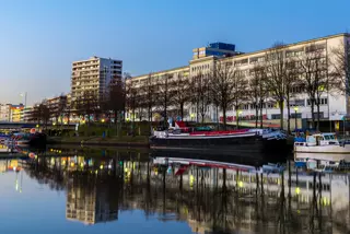 Auf dem Bild ist ein ruhiger Kanal in einer Stadt zu sehen. Am rechten Ufer stehen mehrere mehrstöckige Gebäude, vor denen eine Reihe kahler Bäume verläuft. Entlang des Ufers liegen mehrere Boote und Schiffe vor Anker. Das Wasser ist sehr ruhig und spiegelt die Gebäude, Bäume und Boote nahezu vollständig wider. Der Himmel ist klar und blau, während die Beleuchtung auf die Abendstunden hindeutet. Die Szene vermittelt einen gepflegten und modernen Eindruck einer urbanen Wasserlandschaft.