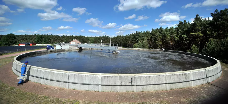 Ein großes, rundes Klärbecken einer Wasseraufbereitungsanlage, umgeben von einem Wald. Im Hintergund der blaue Himmel mit weißen Wolken.