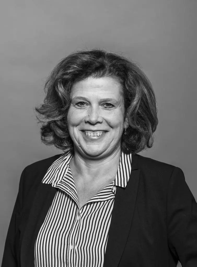 Schwarz-Weiß-Porträt von Kerstin Härtel. Eine lächelnde Frau mittleren Alters mit lockigem Haar. Sie trägt ein gestreiftes Hemd unter einem dunklen Blazer und blickt freundlich in die Kamera. 