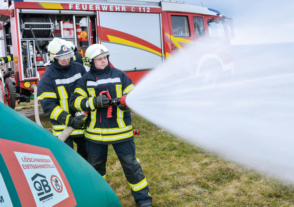 Zwei Feuerwehrmänner stehen vor einem Feuerwehrauto und halten einen Schlauch aus dem Wasser spritzt. Neben ihnen steht eine Löschwasserzisterne