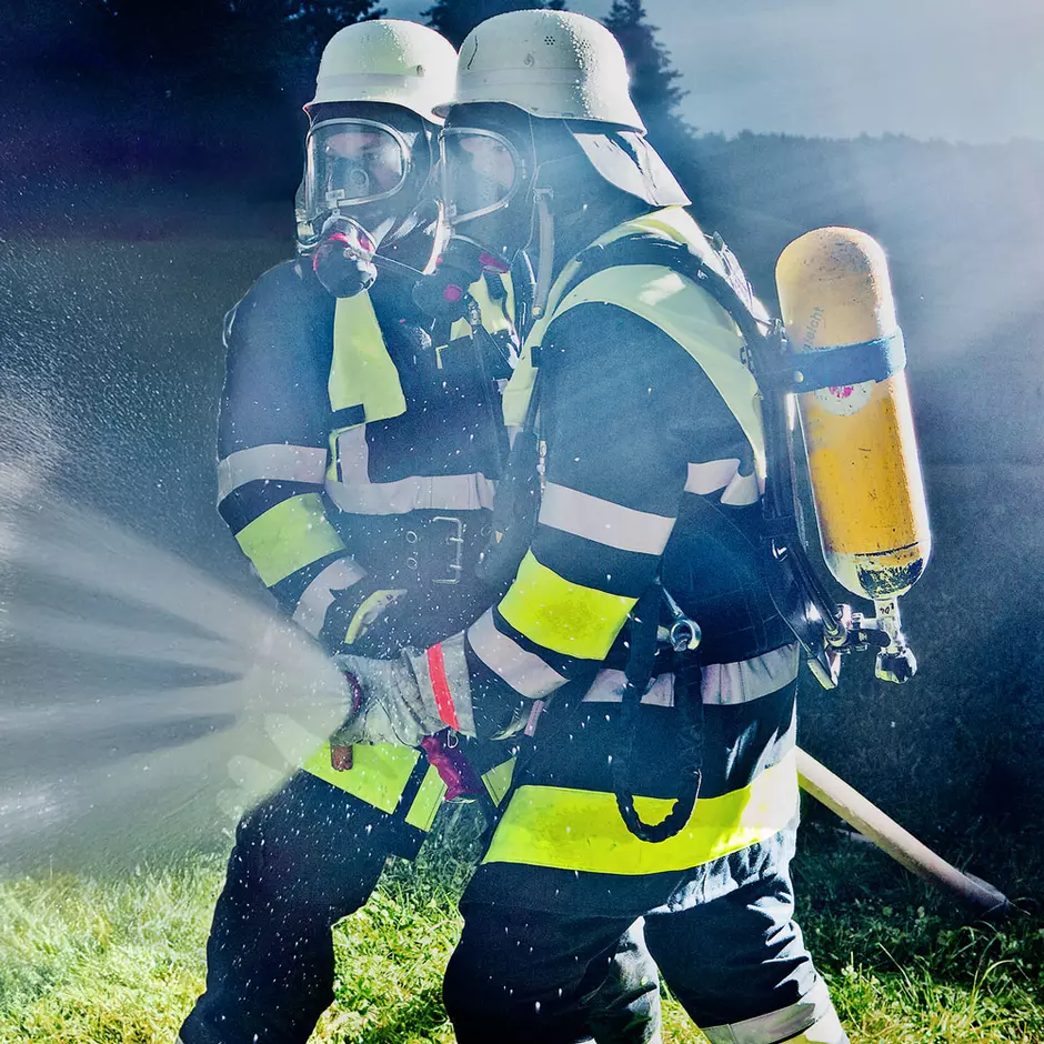 Zwei Feuerwehrmänner stehen vor einem Feuerwehrauto und halten einen Schlauch aus dem Wasser spritzt. Neben ihnen steht eine Löschwasserzisterne
